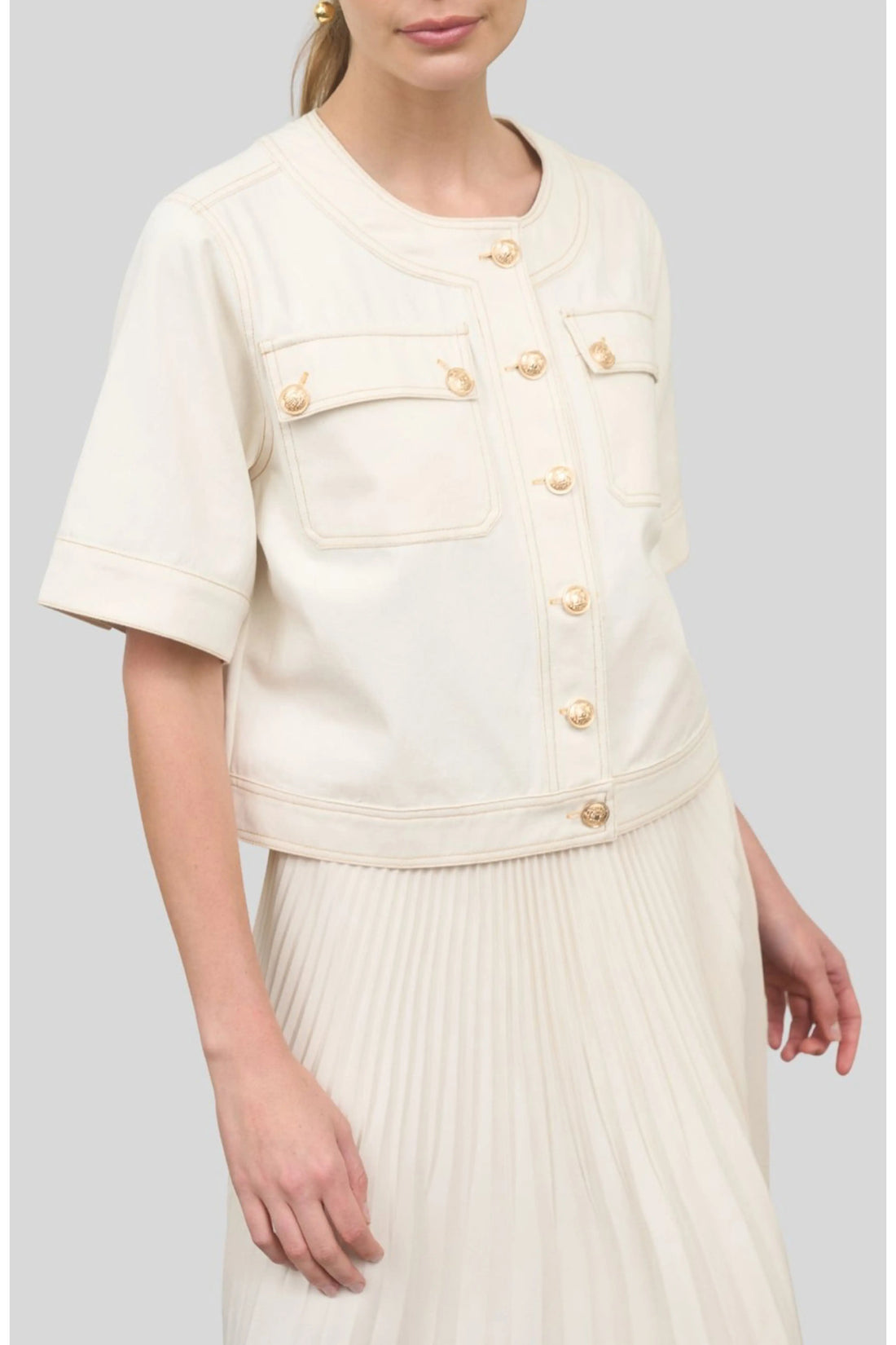 A woman wearing a short-sleeve, collarless cream denim jacket featuring prominent gold crest buttons and chest flap pockets over a pleated skirt.