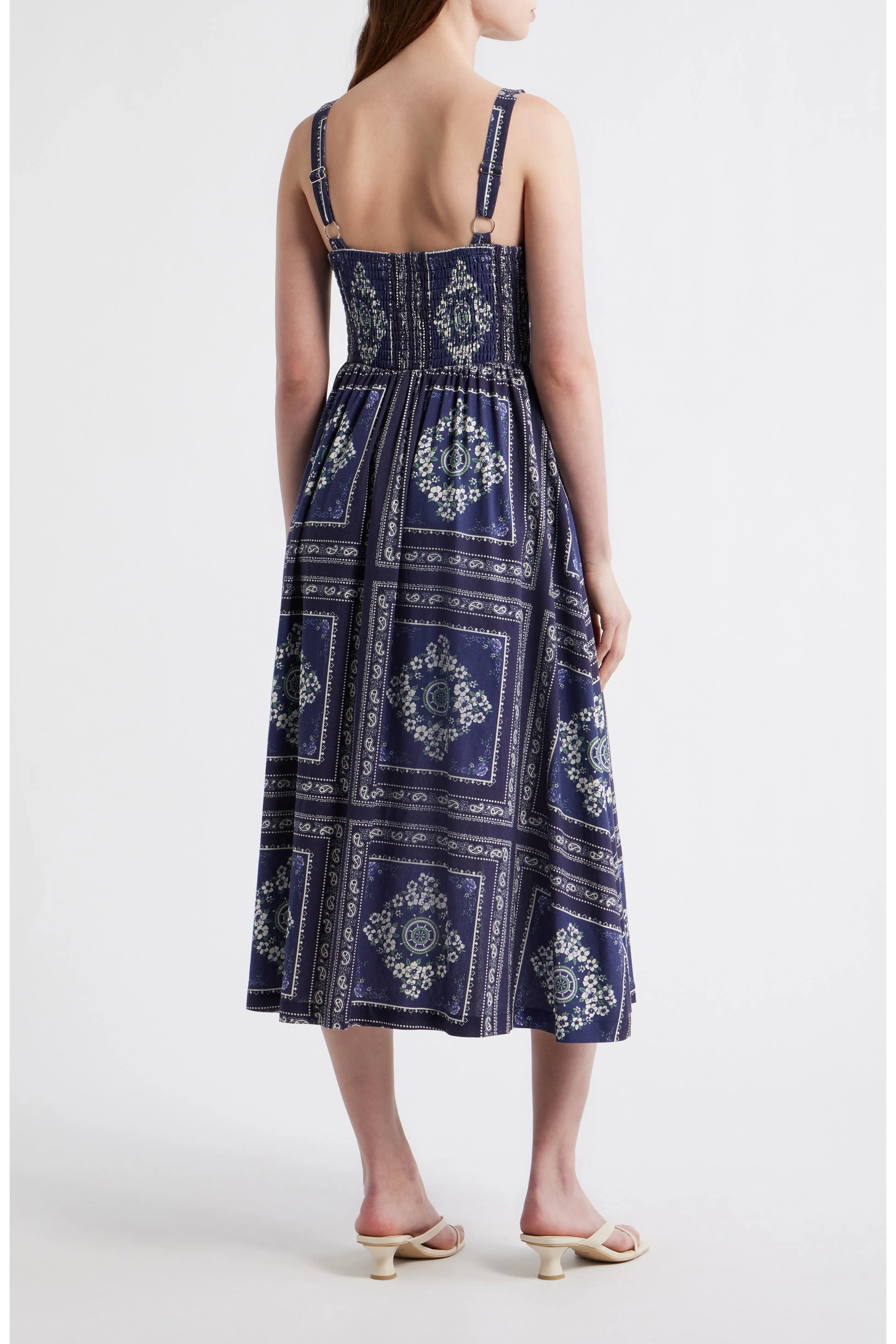 Rear view of a woman wearing a navy blue bandana-patterned cotton-linen midi dress with a square neckline, adjustable straps, and a button-front skirt.