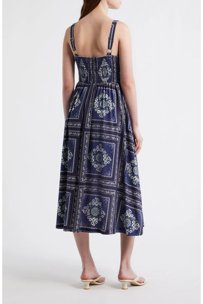 Rear view of a woman wearing a navy blue bandana-patterned cotton-linen midi dress with a square neckline, adjustable straps, and a button-front skirt.