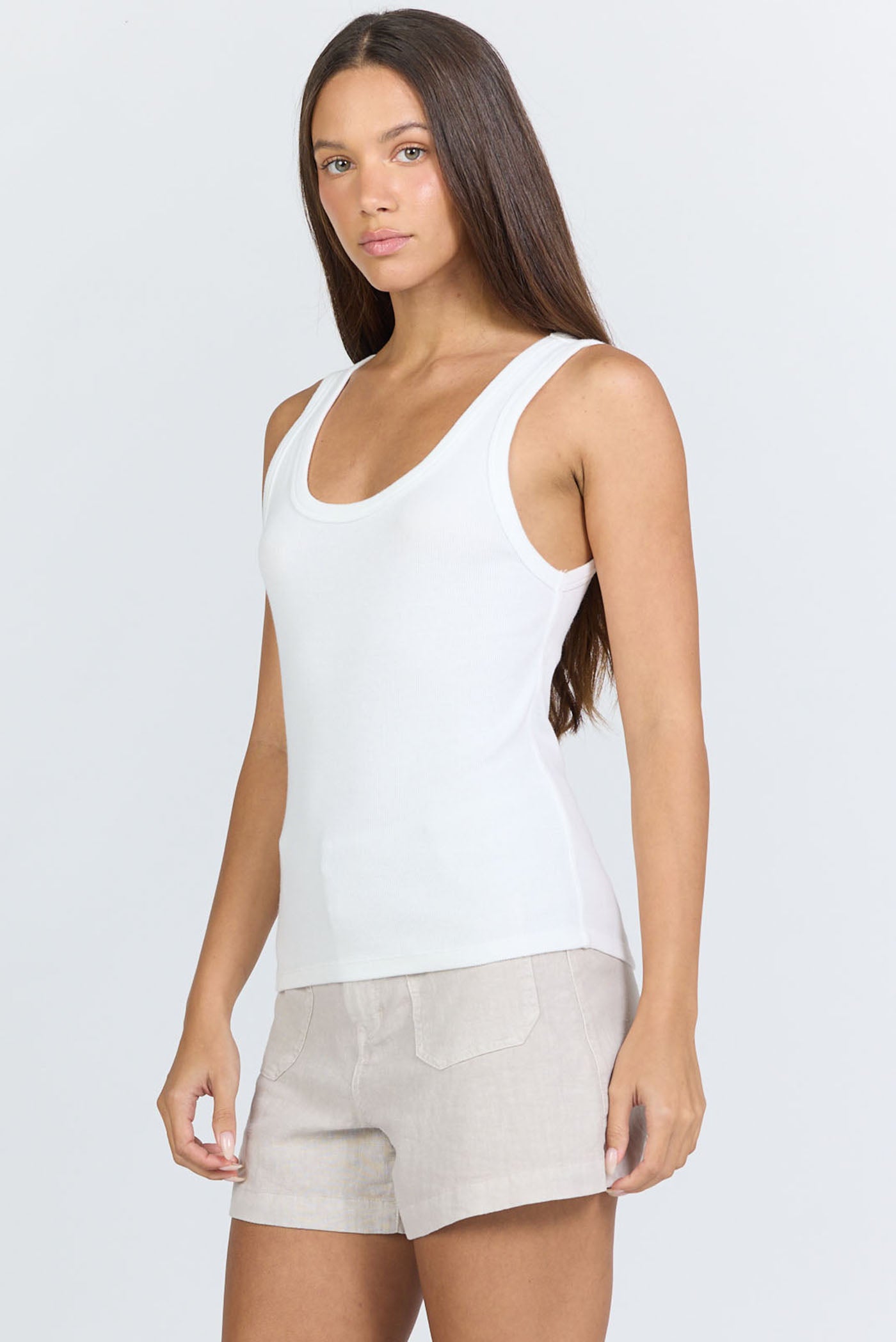 Side view of a woman wearing a fitted white scoop-neck ribbed racerback tank top with beige linen shorts.