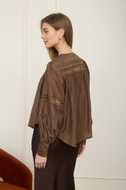 Rear view of a woman wearing a romantic, flowy espresso brown long-sleeve peasant blouse with detailed ladder-lace inserts across the chest and sleeves.