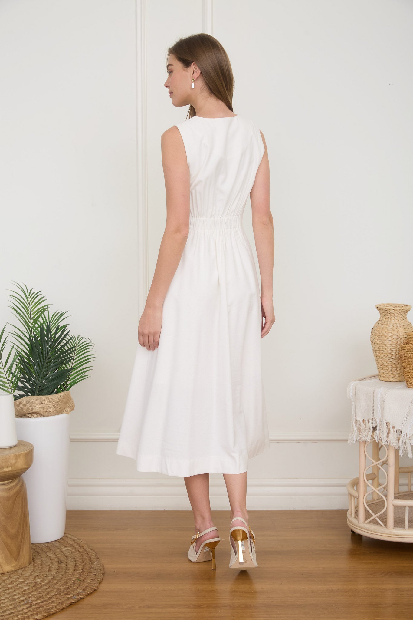 Rear view of a woman wearing a sleeveless, button-front white midi dress with faux hip pockets and low heels.