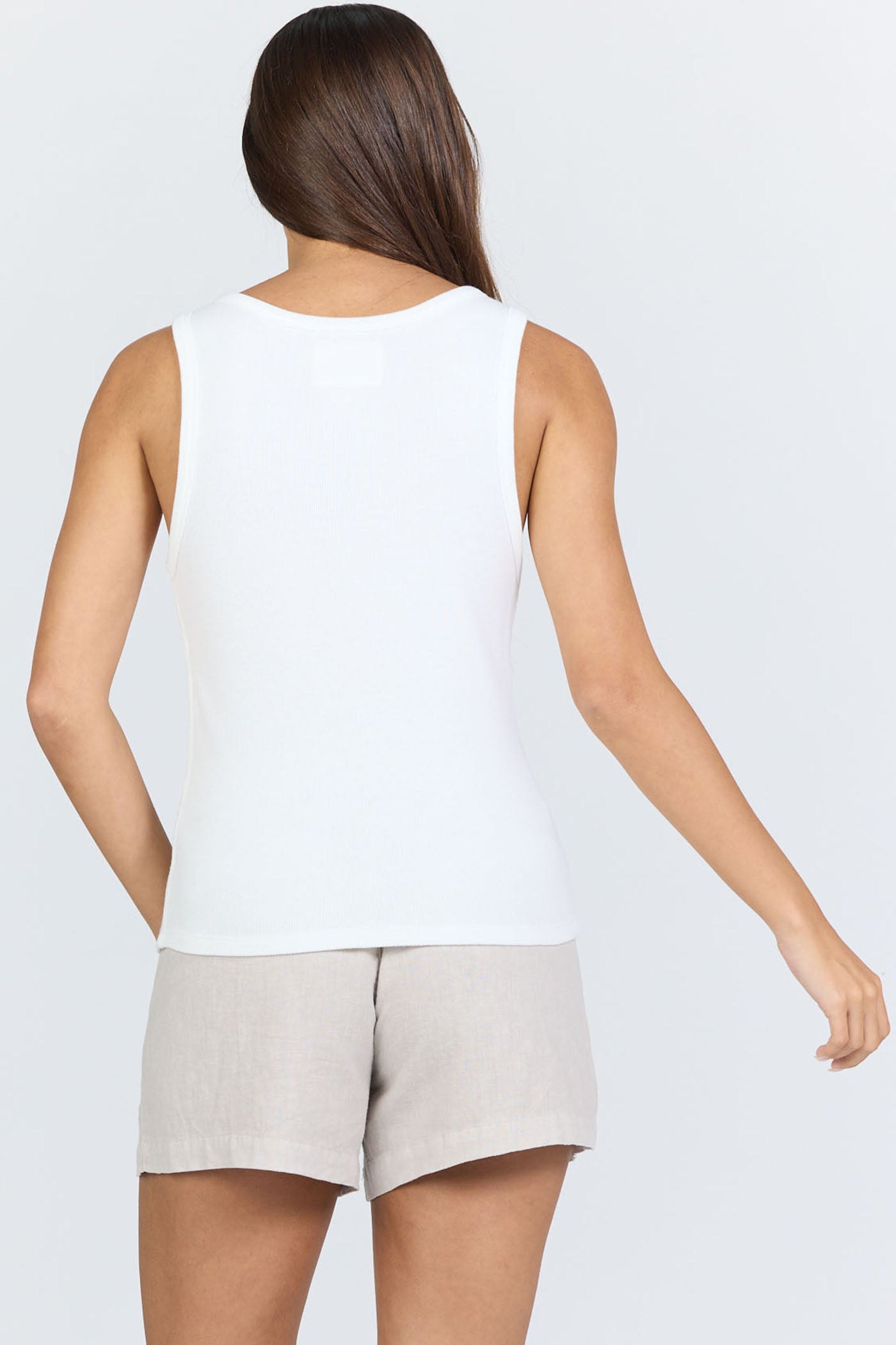 Rear view of a woman wearing a fitted white scoop-neck ribbed racerback tank top with beige linen shorts.