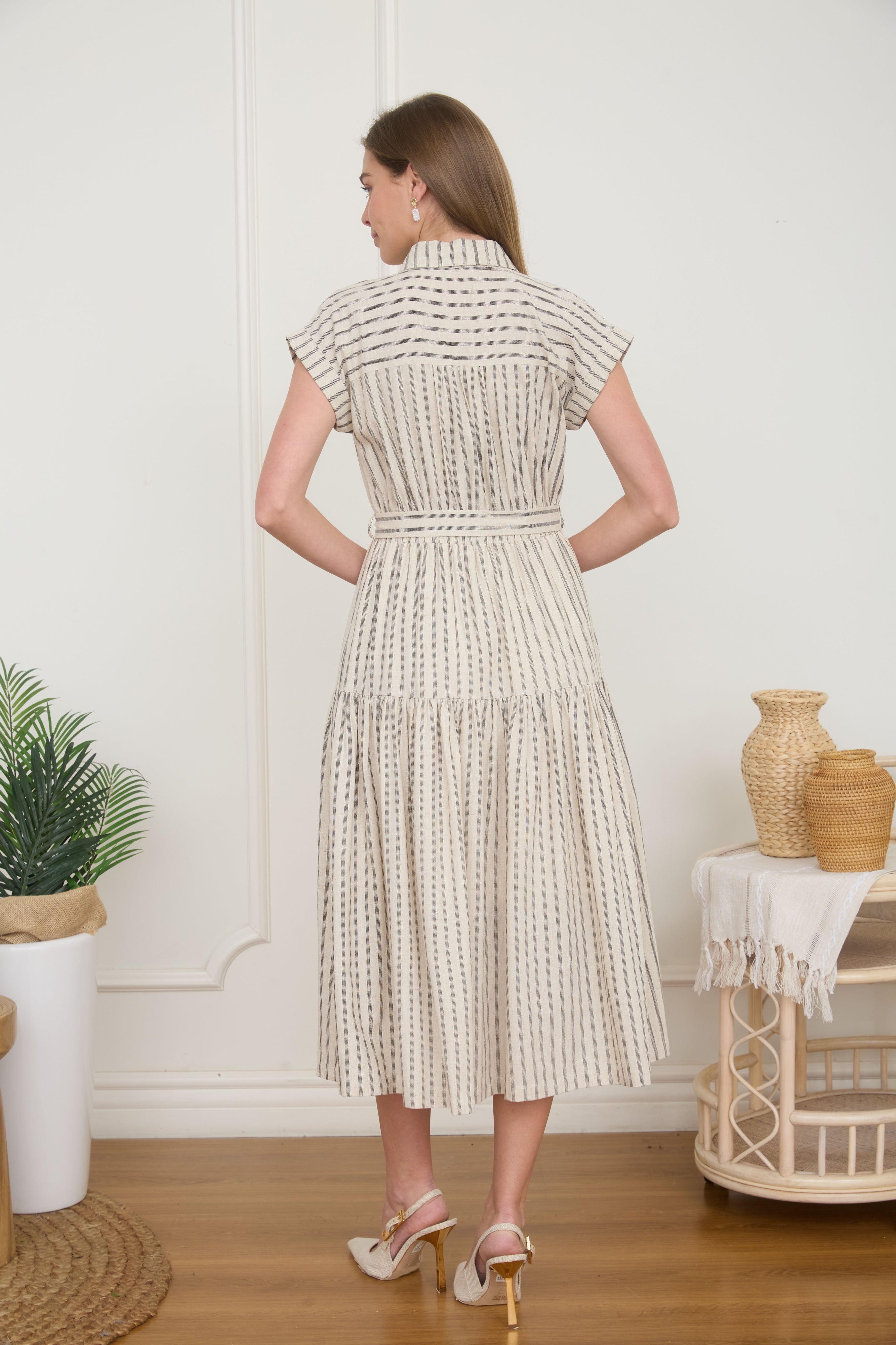 Rear view of a woman wearing a light taupe and cream vertically striped midi shirt dress with a tie belt, short cap sleeves, and high heels.