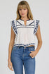 Woman wearing a white sleeveless split-neck top with blue embroidery and blue jeans.