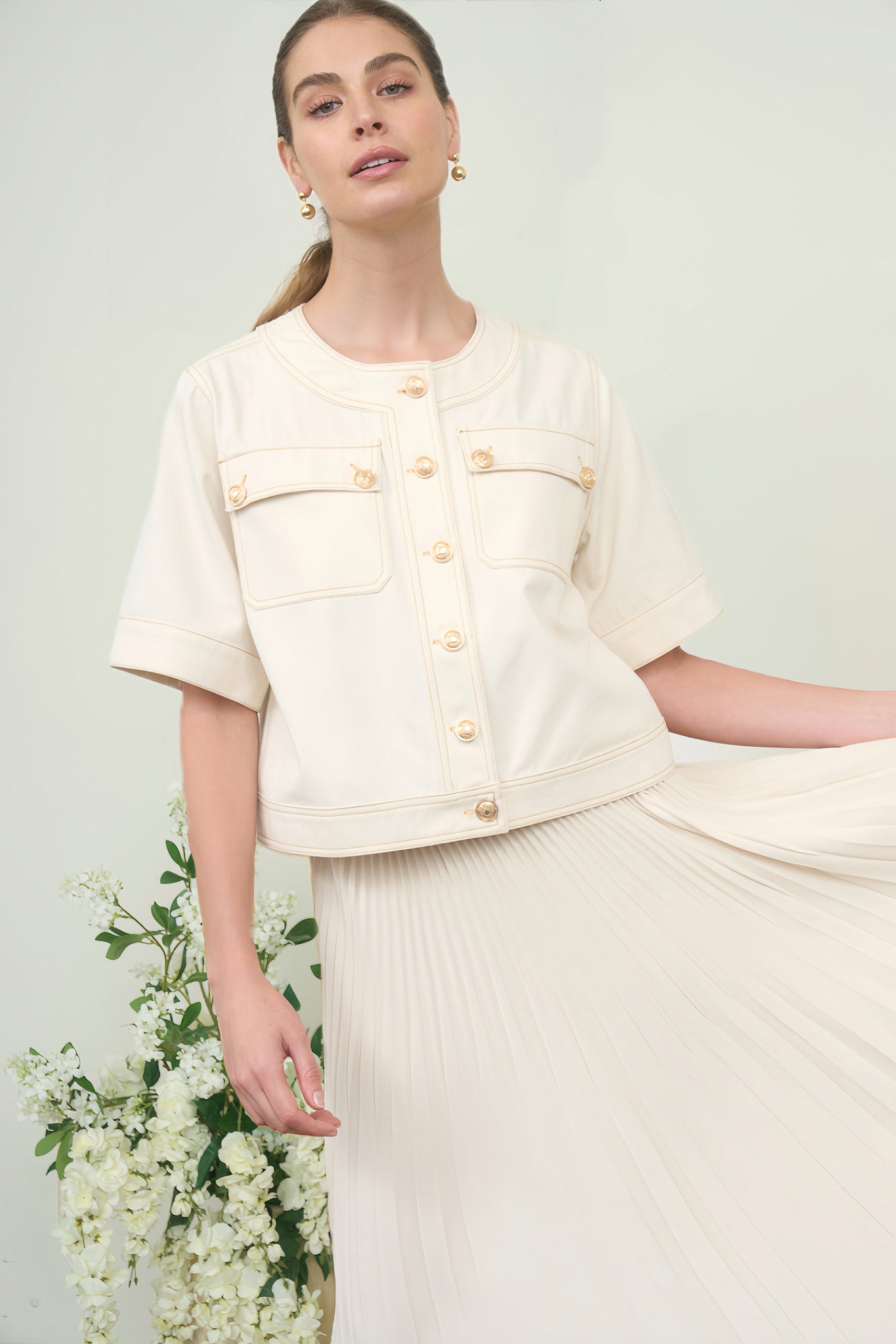 A woman wearing a short-sleeve, collarless cream denim jacket featuring prominent gold crest buttons and chest flap pockets over a pleated skirt.