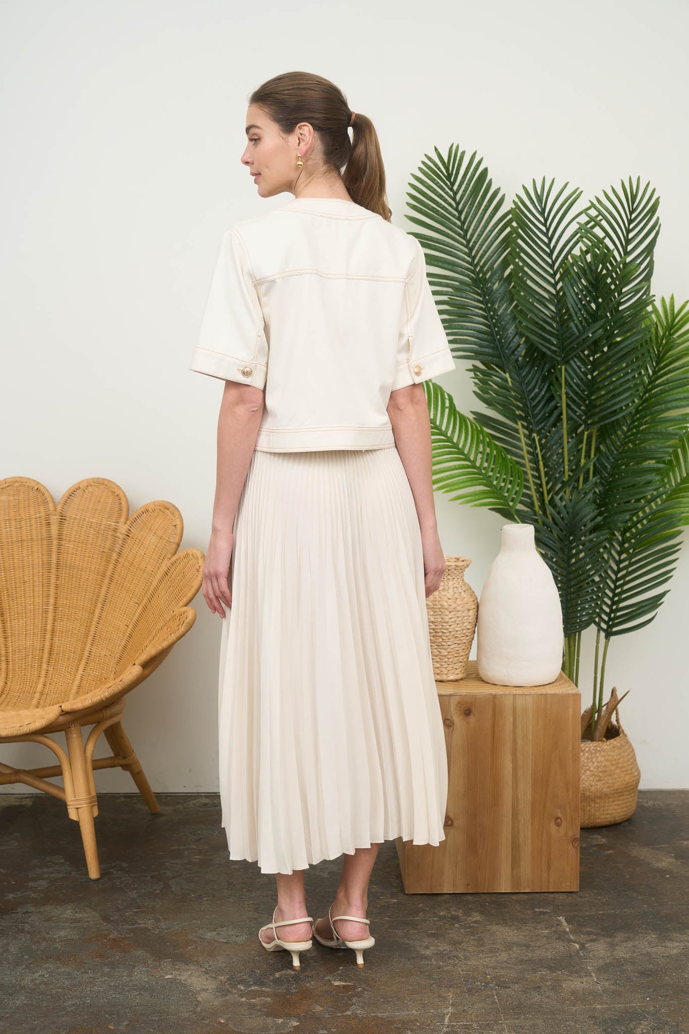 Rear view of a woman wearing a short-sleeve, collarless cream denim jacket over a pleated skirt.