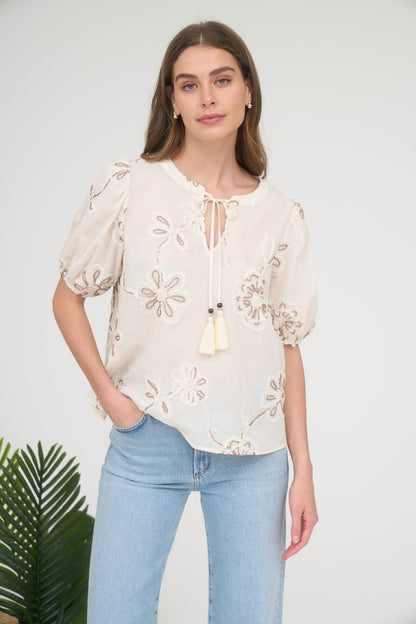 A woman wearing a cream-colored short-sleeve blouse with brown floral embroidery, a split neckline with tassels, and light-wash blue jeans.