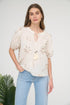 A woman wearing a cream-colored short-sleeve blouse with brown floral embroidery, a split neckline with tassels, and light-wash blue jeans.