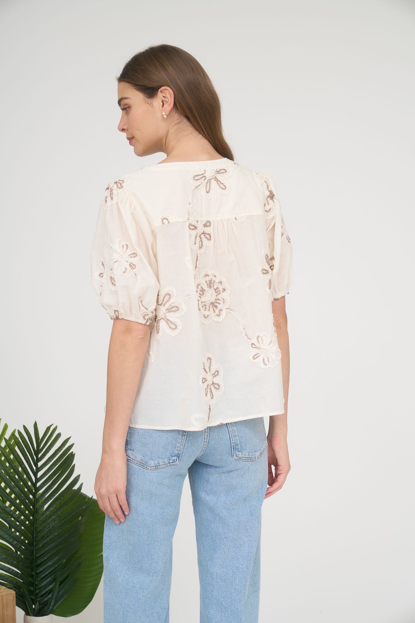 Rear view of a woman wearing a cream-colored short-sleeve blouse with brown floral embroidery and light-wash blue jeans.