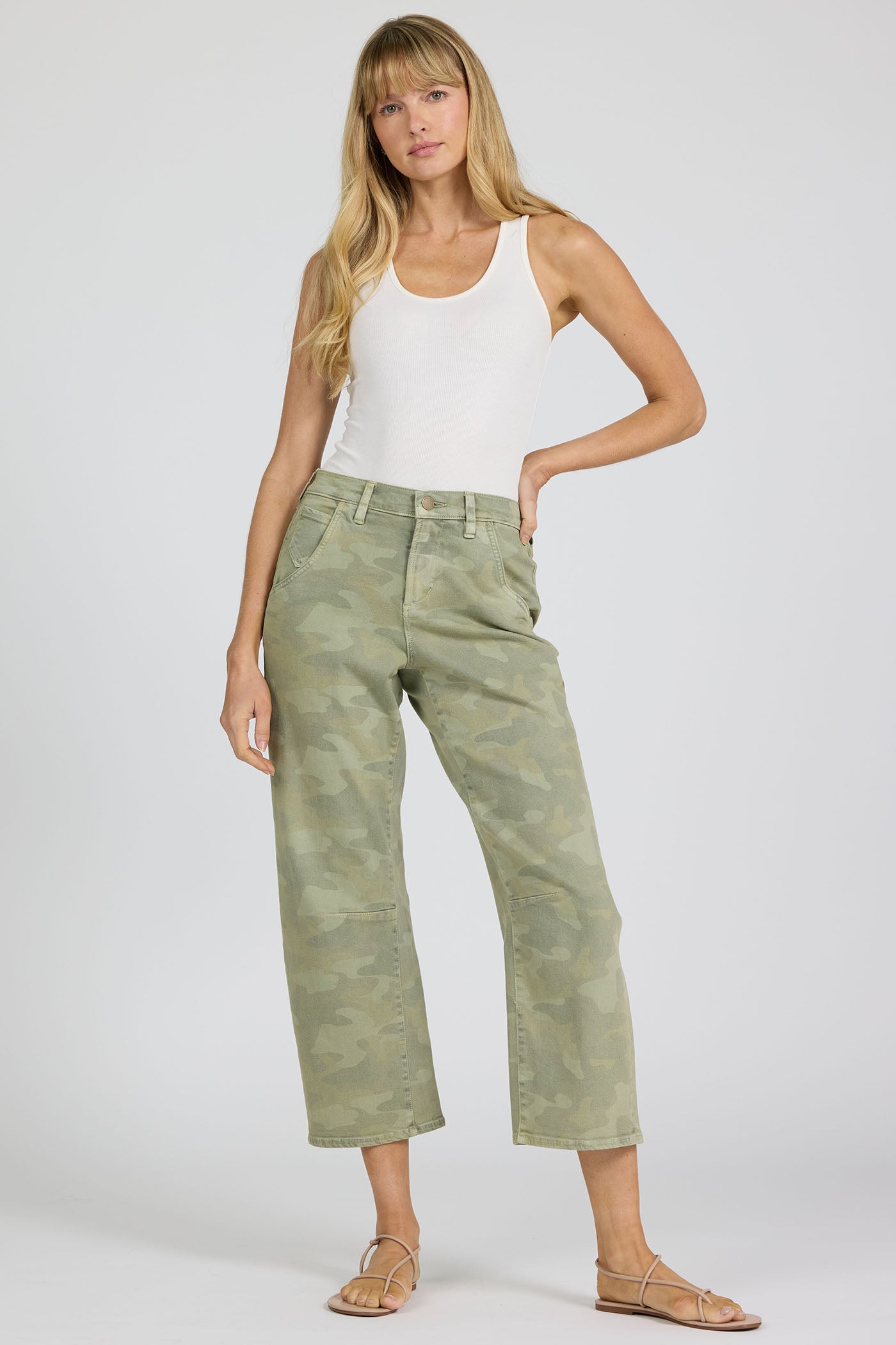 Full body view of a woman wearing green camouflage pants and a white tank top on a white background.