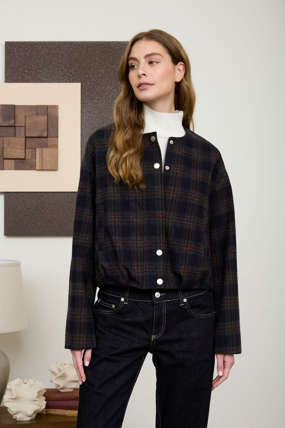 A woman looking to the side wearing a navy and brown plaid collarless bomber jacket layered over a white turtleneck sweater with dark jeans.