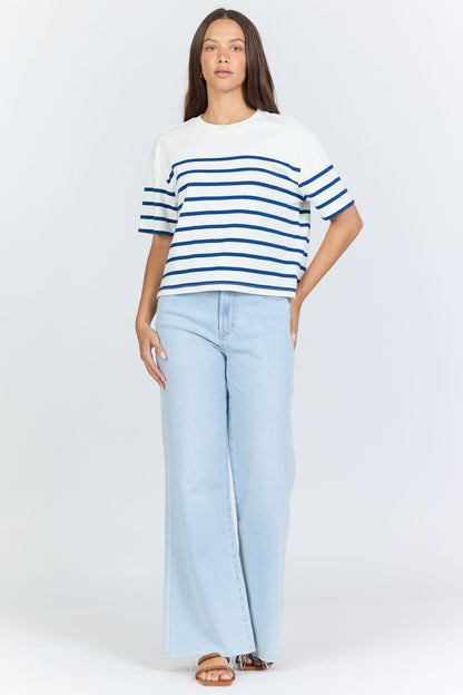 Full body view of a woman wearing a white and blue horizontal striped crew neck cotton t-shirt with light-wash jeans.