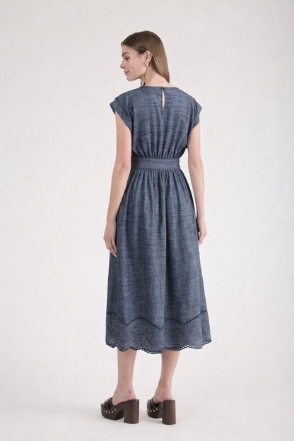 Rear view of a woman wearing a short-sleeve chambray midi dress with a wide grommet belt, a gathered waist, and an intricate floral eyelet scalloped hem.