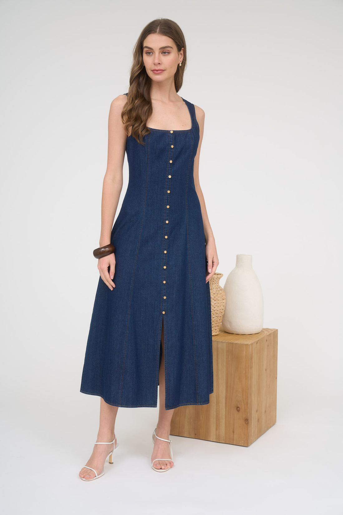 A woman wearing a sleeveless dark wash denim midi dress with a square neckline, copper buttons down the front, and white strappy heels.