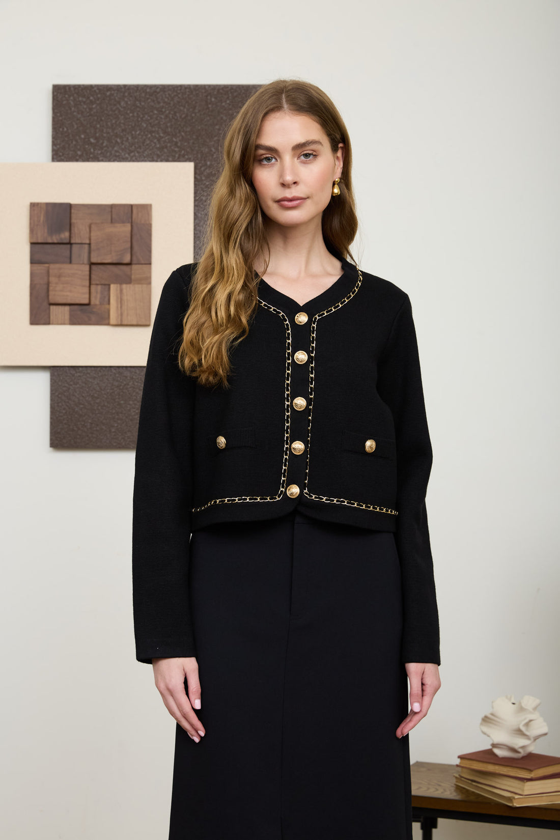 A woman wearing a structured, long-sleeve black knit cardigan featuring a V-neckline, prominent gold crest buttons, and intricate gold chain-link trim.