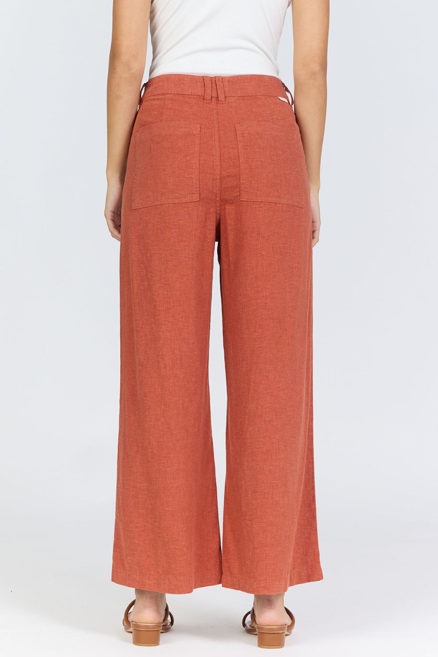 Rear view of a model wearing high-rise linen-blend wide-leg cropped burnt brick colored trousers with a white tucked-in tank top and flat sandals.