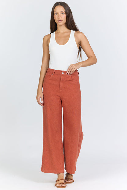 Full body view of a model wearing high-rise linen-blend wide-leg cropped burnt brick colored trousers with a white tucked-in tank top and flat sandals.