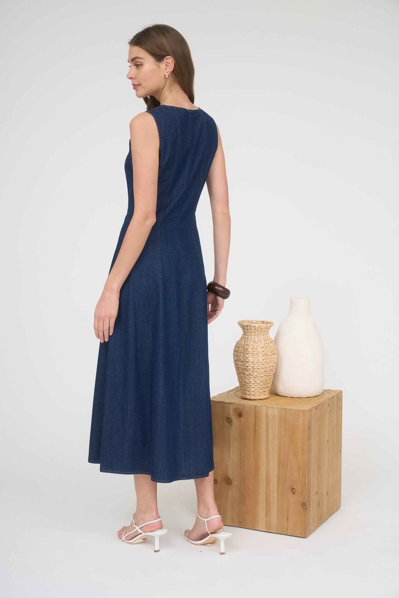 Rear view of a woman wearing a sleeveless dark wash denim midi dress and white strappy heels.