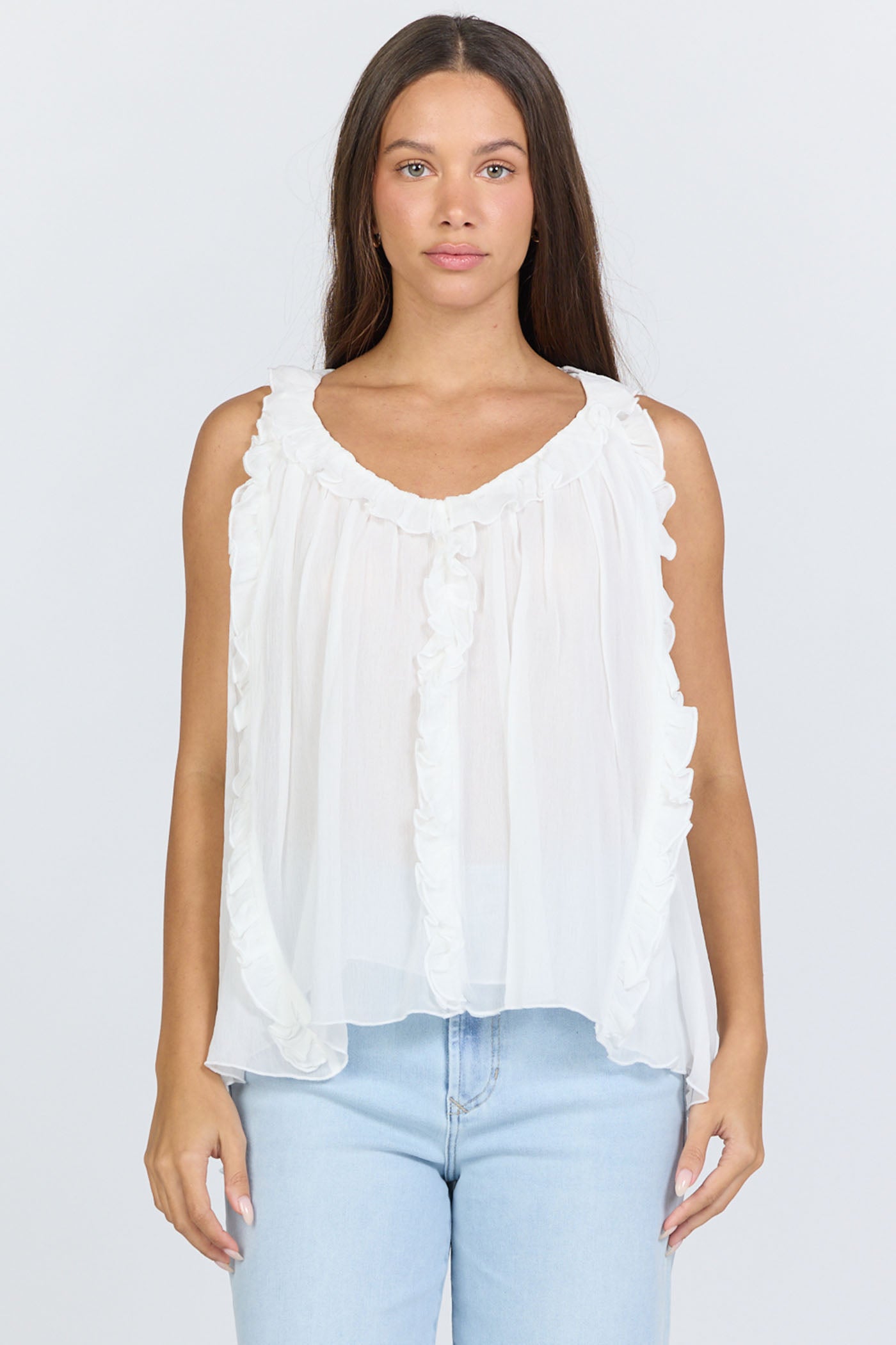 Front view of a woman wearing a flowy white sleeveless scoop-neck top with vertical ruffles and light blue jeans.