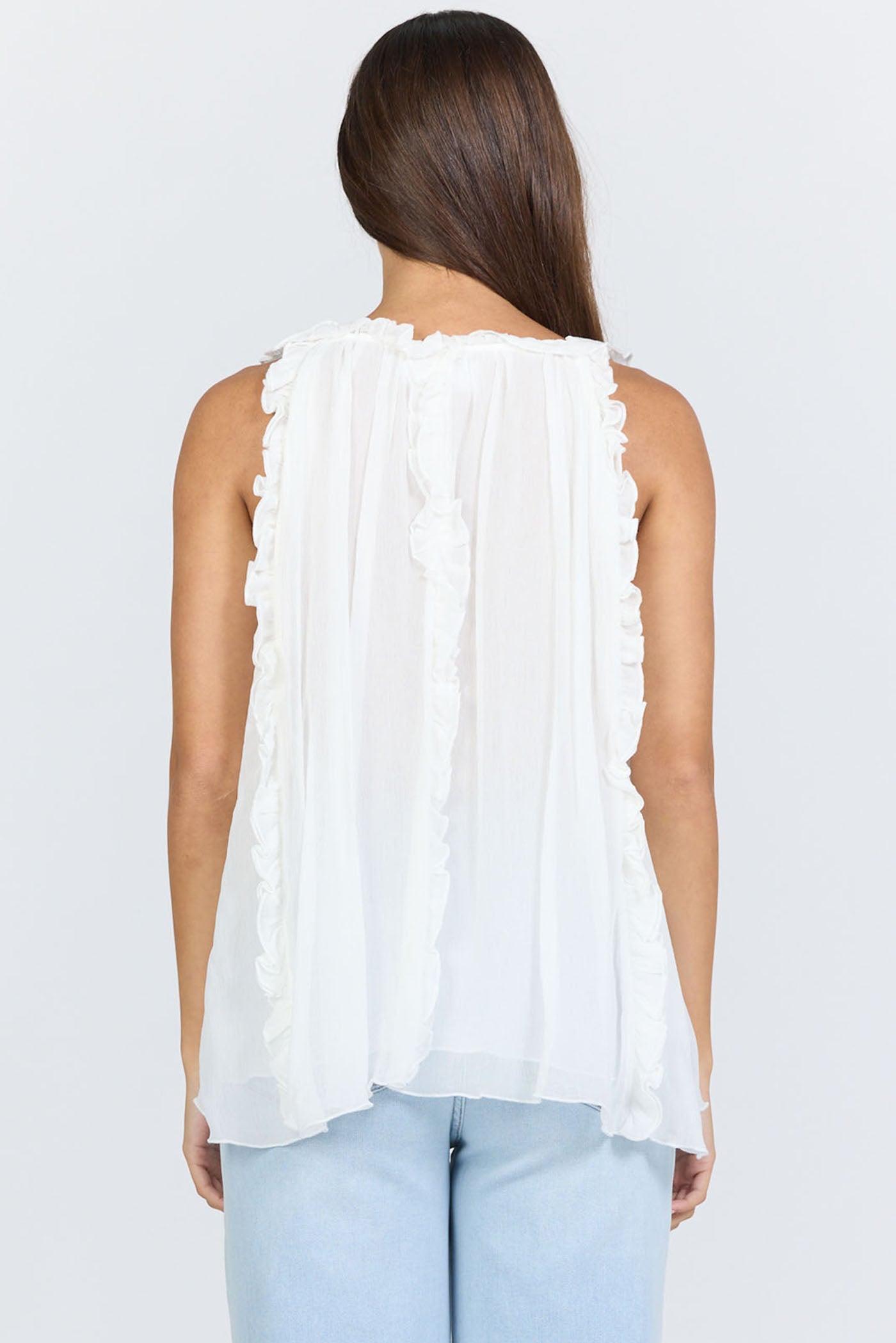 Rear view of a woman wearing a flowy white sleeveless scoop-neck top with vertical ruffles and light blue jeans.