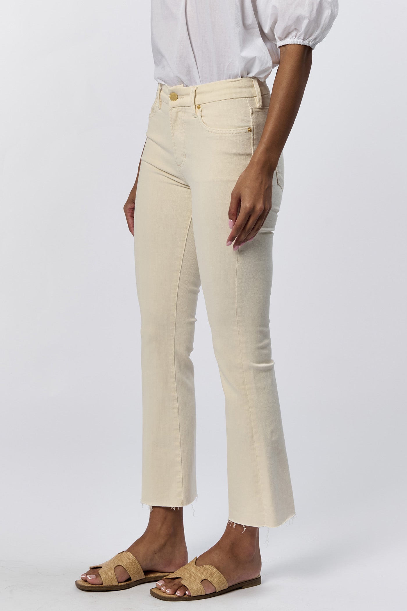 Side view of a woman wearing the cream mid-rise raw hem flare jeans with gold hardware and a white top.