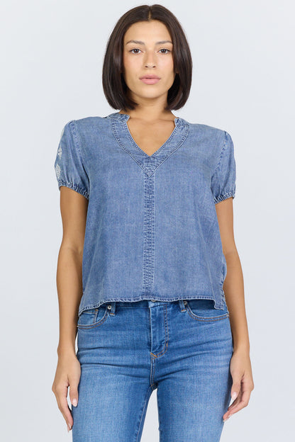 Front view of a woman wearing a light blue 100% Tencel V-neck blouse with embroidered short puff sleeves, paired with denim jeans.