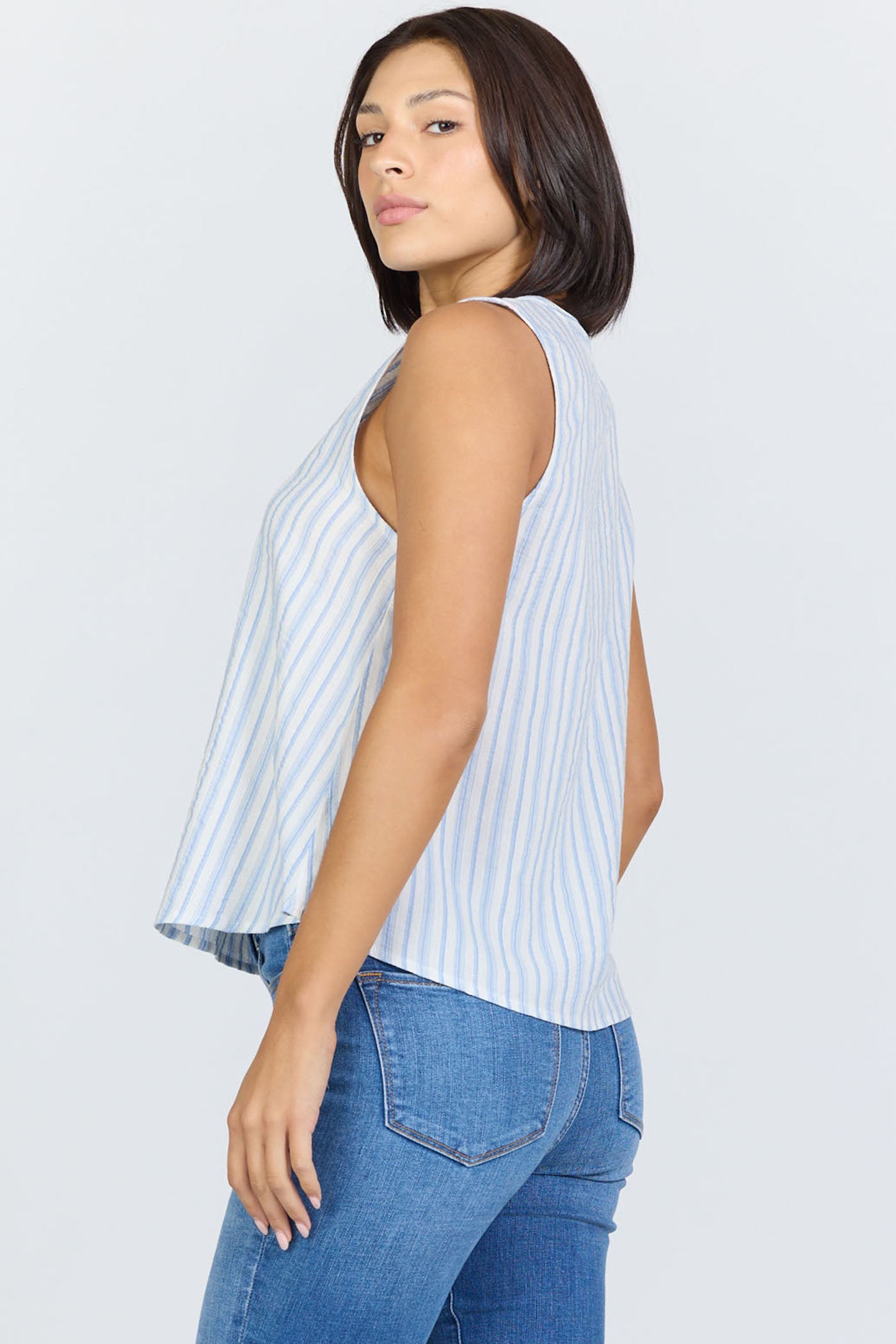 Side view of a woman with a dark bob haircut wearing a sleeveless blue and white striped cotton-blend top with a button-up Henley neck and blue jeans.