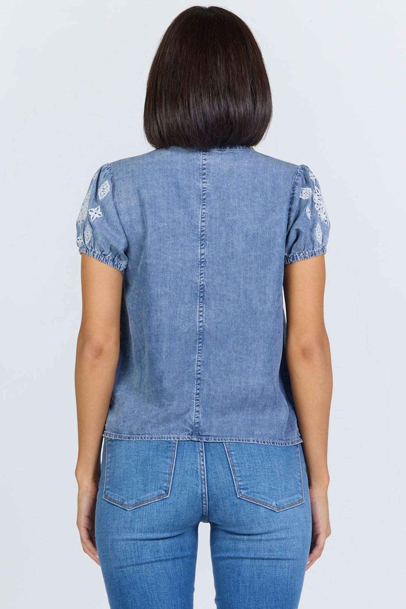 Rear view of a woman wearing a light blue 100% Tencel V-neck blouse with embroidered short puff sleeves, paired with denim jeans.