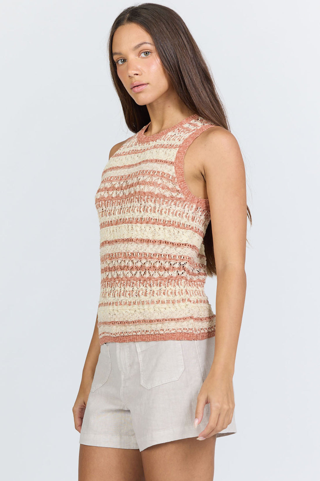 Side view of a woman wearing a sleeveless crew-neck sweater tank with terra-cotta and ivory horizontal stripes, paired with light linen shorts.