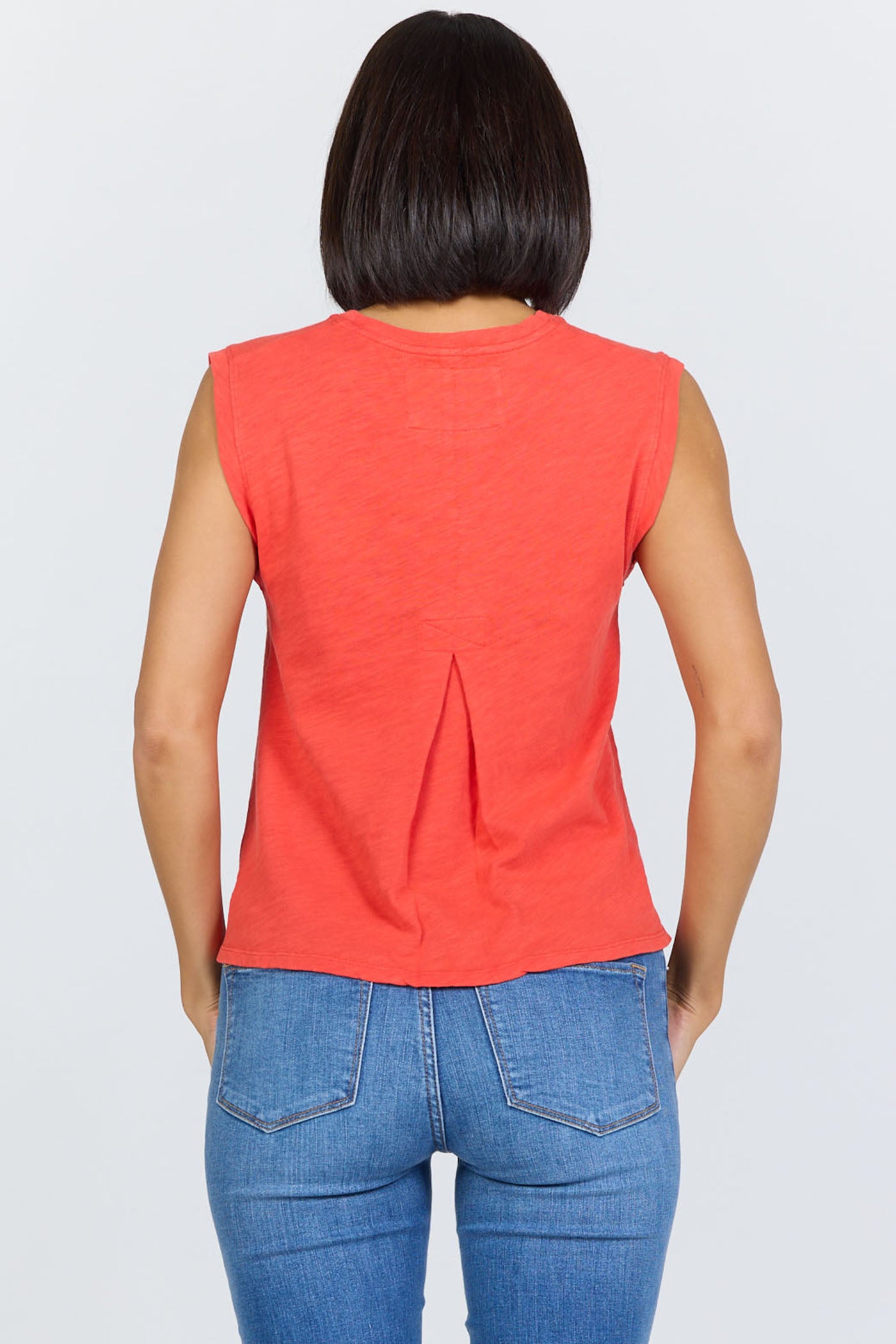 Rear view of a woman wearing a poppy red sleeveless crew neck muscle tank made from textured slub cotton, paired with blue jeans.