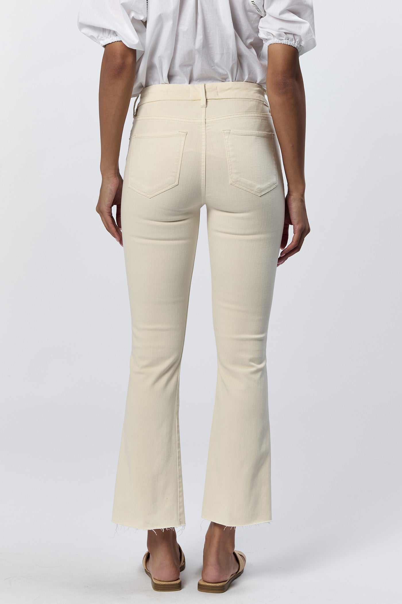 Rear view of a woman wearing the cream mid-rise raw hem flare jeans with gold hardware and a white top.