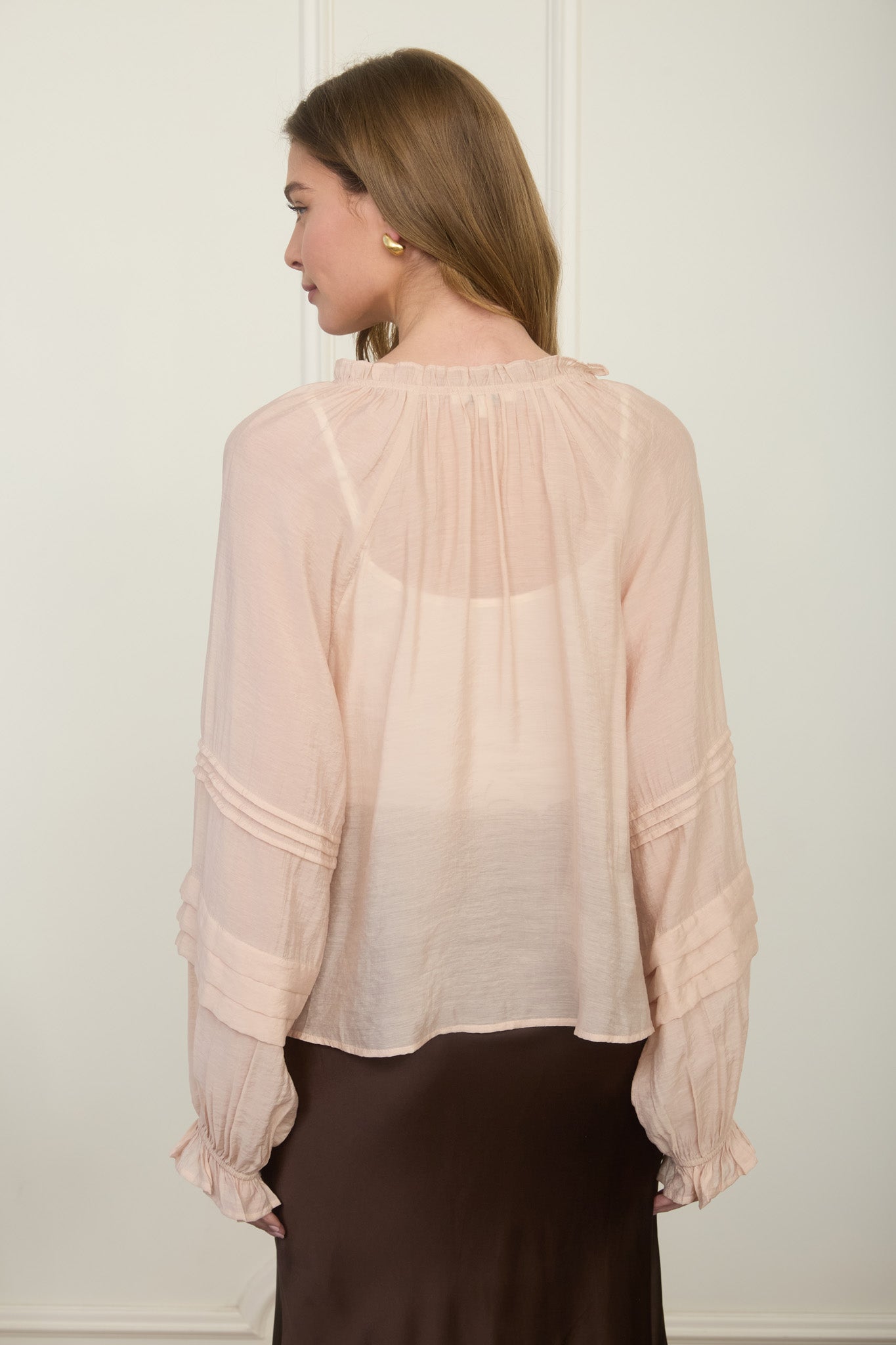 Rear view of a woman wearing a romantic, flowy blush pink long-sleeve blouse featuring intricate tiered pleats on the sleeves.