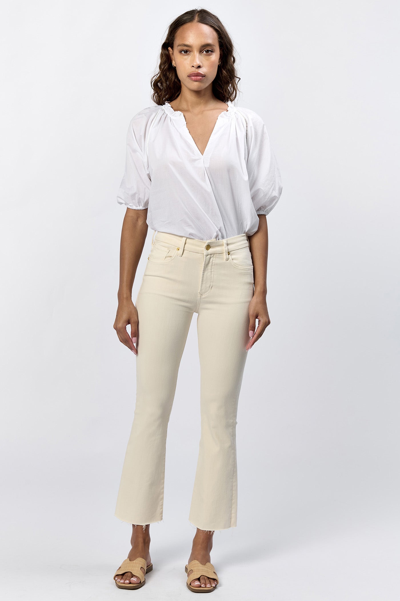 Full body view of a woman wearing the cream mid-rise raw hem flare jeans with gold hardware and a white top