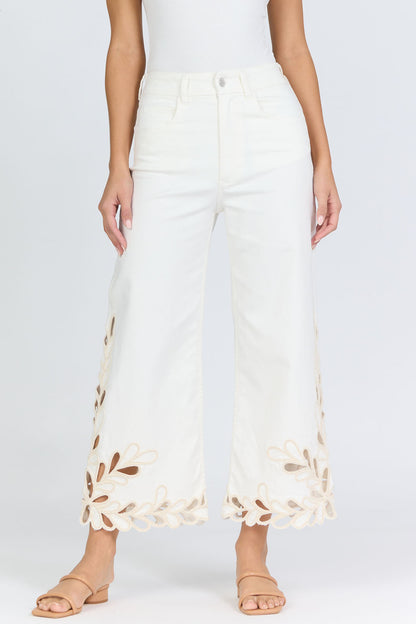 Front view of a woman wearing high-rise white wide-leg cropped jeans with an intricate floral laser-cut scalloped hemline and tan slide sandals.