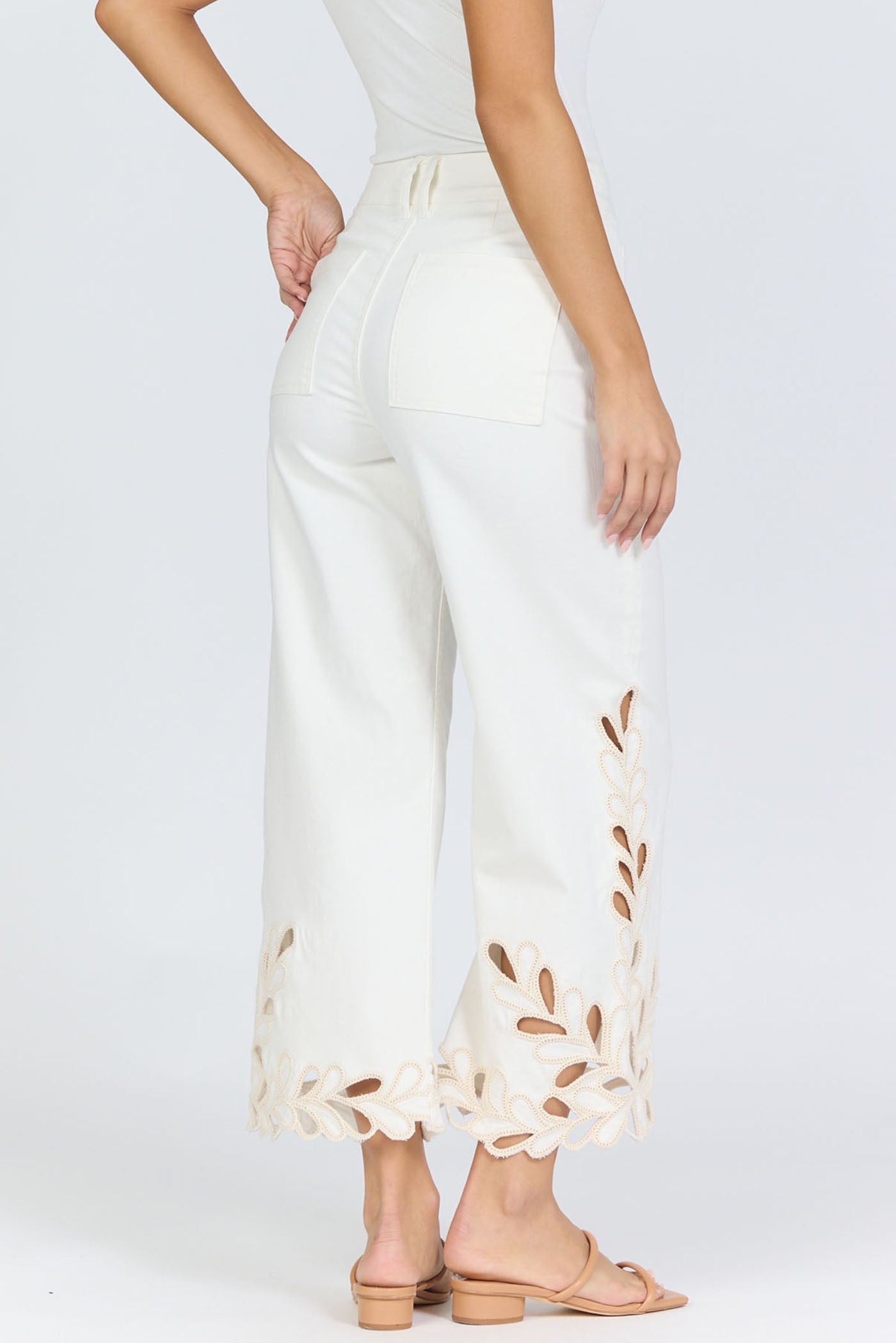 Rear view of a woman wearing high-rise white wide-leg cropped jeans with an intricate floral laser-cut scalloped hemline and tan slide sandals.