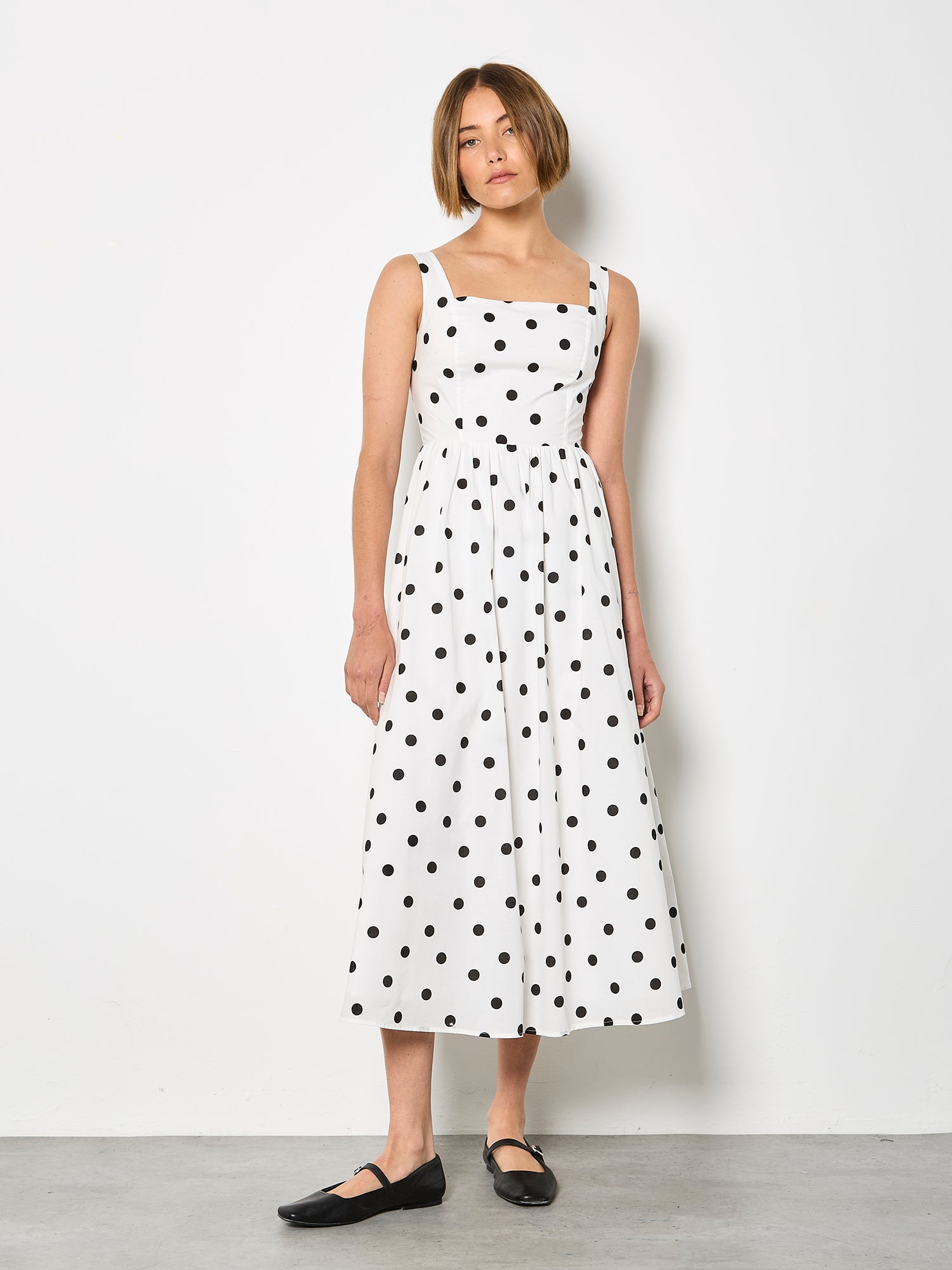 Front view of a model with short brown hair wearing a white midi dress with large black polka dots and black flats.