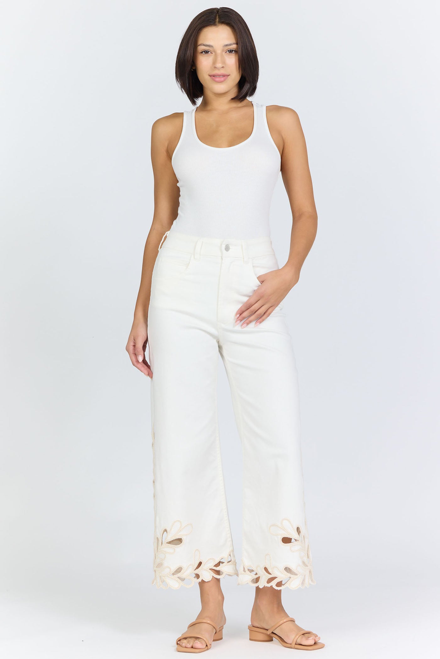Full body view of a woman wearing high-rise white wide-leg cropped jeans with an intricate floral laser-cut scalloped hemline and tan slide sandals.