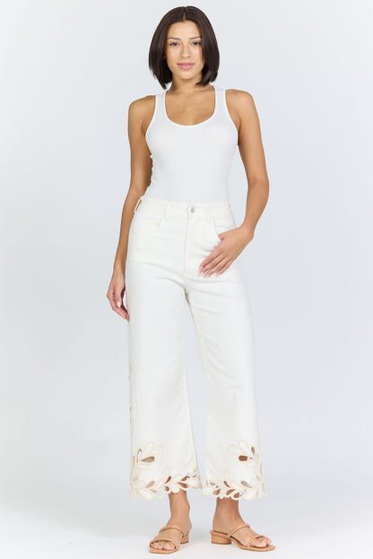 Full body view of a woman wearing high-rise white wide-leg cropped jeans with an intricate floral laser-cut scalloped hemline and tan slide sandals.