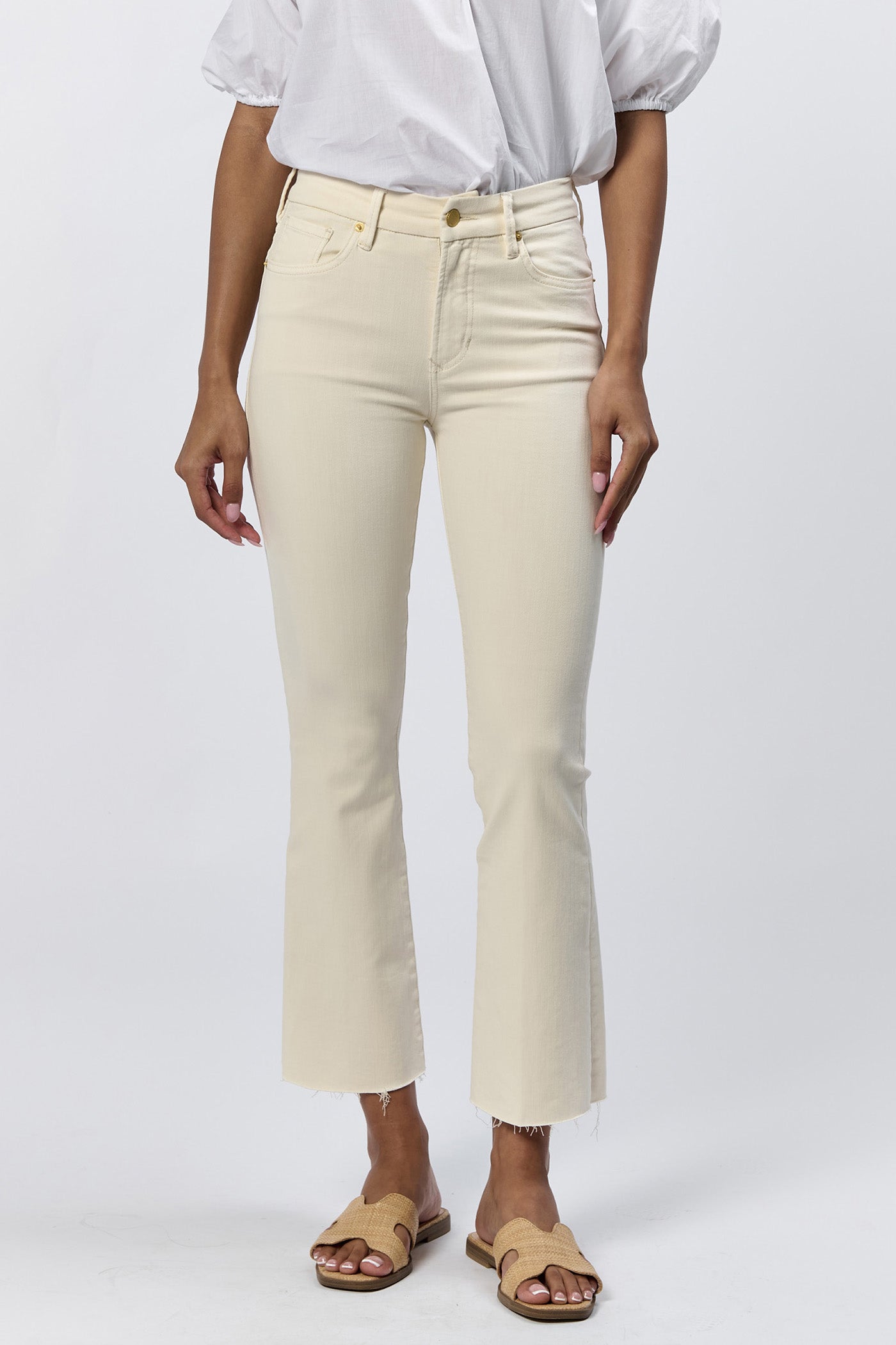 Front view of a woman wearing the cream mid-rise raw hem flare jeans with gold hardware and a white top.